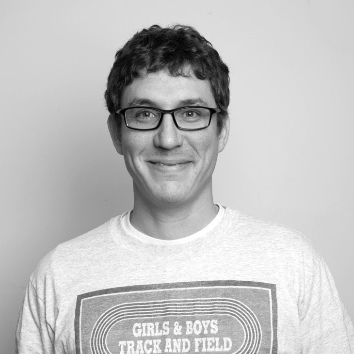 A Caucasian male with short curly brown hair, wearing glasses and a white t-shirt with "GIRLS & BOYS TRACK AND FIELD" printed on it, smiling against a gray background.
